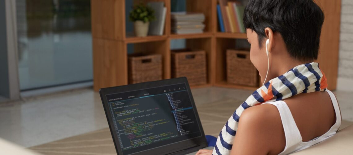 Eine kurzhaarige Frau von hinten gesehen, die an einem Laptop arbeitet, auf dessen Bildschirm Code zu sehen ist, in einem modernen Wohnzimmer mit Bücherregalen und Körben im Hintergrund.